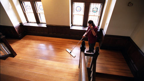 Custodian mops gleaming wood floor as sun pours through stained glass windows
