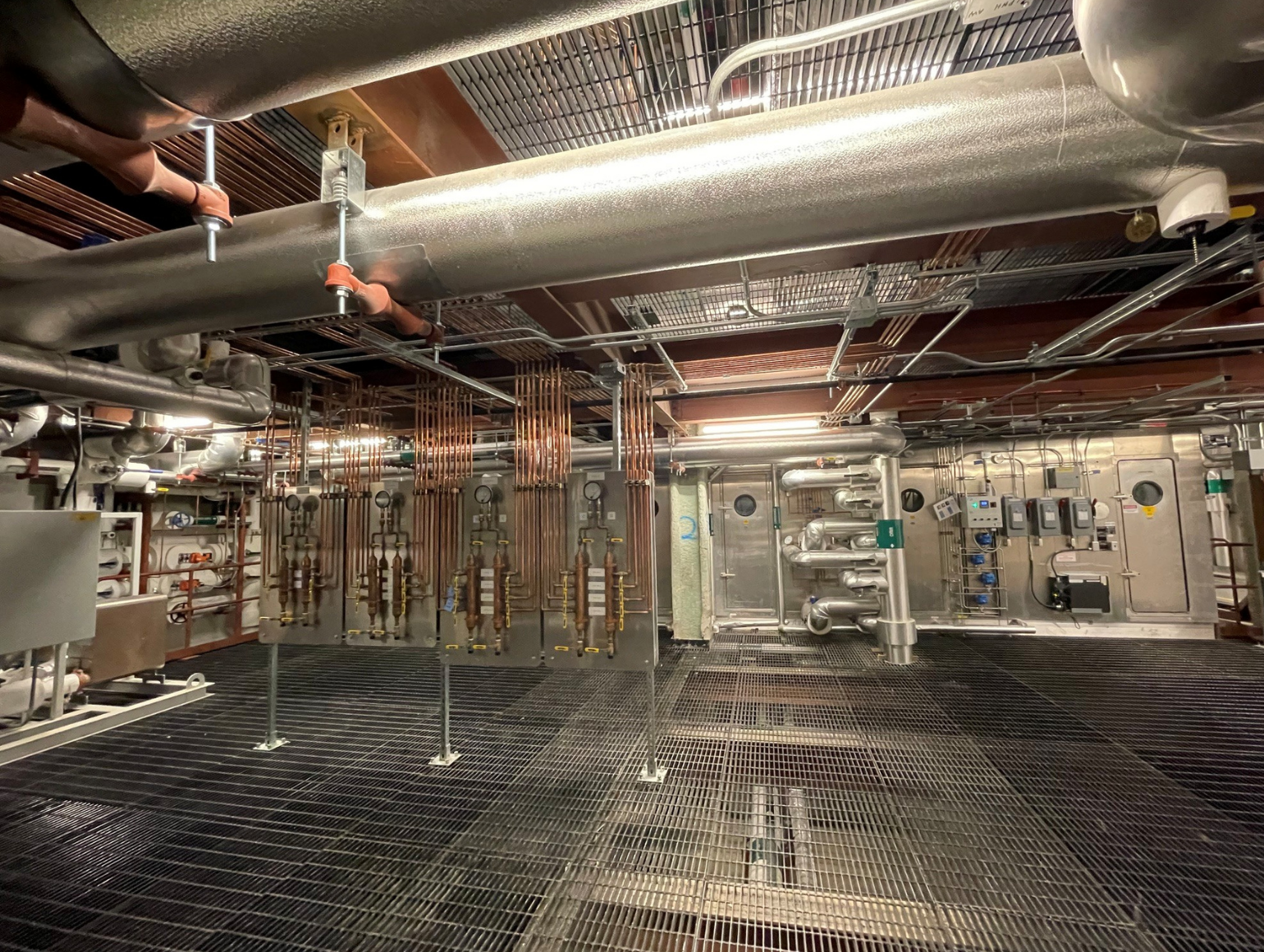 Bright mechanical room with intricate piping, overhead ducts and floor grates